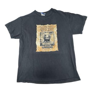 May include: Black t-shirt featuring a distressed poster design. The poster reads "Have You Seen This Wizard?" with an image of a man. The shirt is a crew neck style.