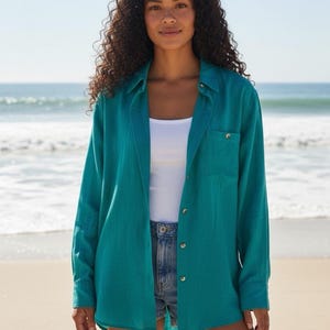 May include: Teal long-sleeved shirt with a button-down front and a chest pocket. The shirt is worn over a white vest top and paired with denim shorts. The model is standing on a sandy beach with the sea in the background.