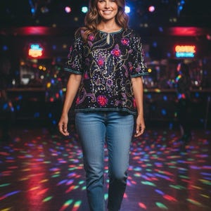May include: A woman wearing a black sequined top with colorful floral embroidery, paired with blue jeans and white sneakers. The top features a keyhole neckline and short sleeves. The background shows a disco ball and colorful lights.