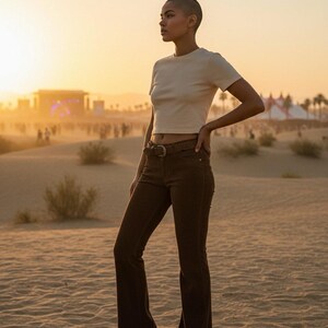 May include: A person wearing a cream-colored, short-sleeved crop top and brown flared pants with a woven belt. The person is standing in a desert landscape at sunset. The pants have a high waist and a flared leg.