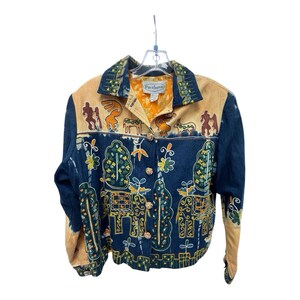 May include: A denim jacket with tan suede accents and intricate embroidery. The jacket features a button-down front, a collared neckline, and long sleeves. The embroidery includes figures, trees, and floral designs in shades of yellow, green, and orange.