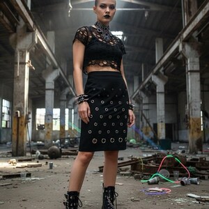 May include: A black outfit featuring a lace crop top, a skirt with metal eyelets, and platform boots with chain details. The model is wearing a choker and spiked bracelets. The setting is an industrial space.