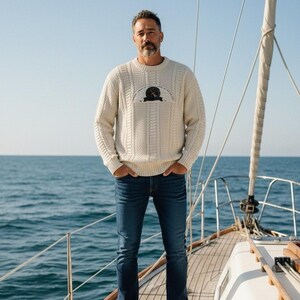 Op de afbeelding: Een man draagt een crèmekleurige kabelgebreide trui met een grafische print op de borst, gecombineerd met een donkerblauwe jeans en witte sneakers. Hij staat op het dek van een boot met de zee op de achtergrond.