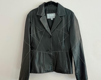 May include: Black leather jacket with geometric paneling and white stitching. The jacket features a notched collar, two-button closure, and a tailored fit. A small label is visible near the collar. The jacket is hanging on a black hanger.