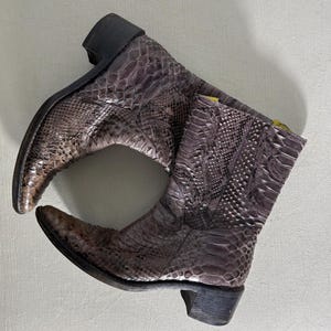 May include: A pair of cowboy boots with a snakeskin pattern in shades of brown and gray. The boots have a pointed toe, a short heel, and a pull-on design. The boots are made of a textured material.