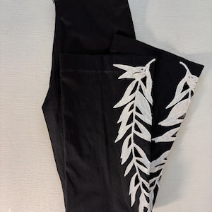 Puede incluir: Pantalones negros con bordado de hojas blancas en el lateral. Los pantalones están doblados, mostrando el intrincado diseño de hojas blancas sobre la tela oscura. Los pantalones tienen una cremallera.