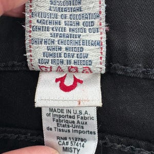 Puede incluir: Primer plano de una etiqueta de prenda negra con texto blanco. La etiqueta incluye instrucciones de lavado, composici&oacute;n de la tela y el texto "MADE IN U.S.A.". El logotipo de la marca es una herradura roja. La palabra "MISTY" tambi&eacute;n es visible.