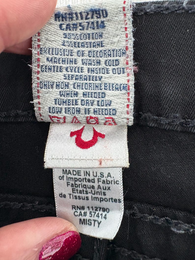 Puede incluir: Primer plano de una etiqueta de prenda negra con texto blanco. La etiqueta incluye instrucciones de lavado, composici&oacute;n de la tela y el texto "MADE IN U.S.A.". El logotipo de la marca es una herradura roja. La palabra "MISTY" tambi&eacute;n es visible.