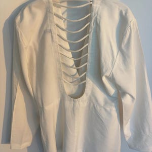 Puede incluir: Camisa blanca de manga larga con un dise&ntilde;o de espalda abierta &uacute;nico. La espalda presenta una serie de cortes verticales paralelos, creando un efecto de celos&iacute;a. La camisa tiene un cuello cl&aacute;sico y pu&ntilde;os abotonados.