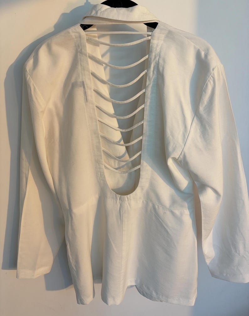 Puede incluir: Camisa blanca de manga larga con un dise&ntilde;o de espalda abierta &uacute;nico. La espalda presenta una serie de cortes verticales paralelos, creando un efecto de celos&iacute;a. La camisa tiene un cuello cl&aacute;sico y pu&ntilde;os abotonados.