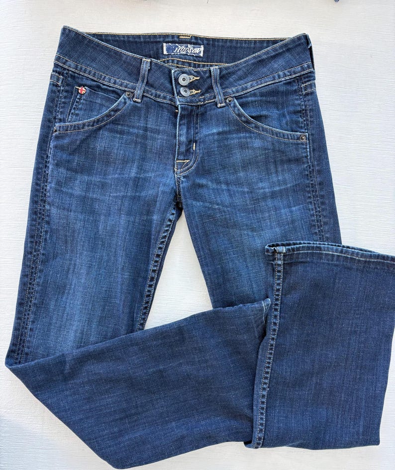 Może przedstawiać: Ciemnoniebieskie dżinsy z klasycznym pięciokieszeniowym designem. Posiadają zapięcie na guzik i zamek błyskawiczny oraz mały czerwony emblemat na przedniej kieszeni. Dżinsy są złożone, prezentując tylne kieszenie i metkę marki.