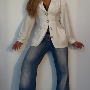 Puede incluir: Una blazer blanca con tres botones negros y dos bolsillos, combinada con vaqueros azules. La blazer tiene un corte entallado y los vaqueros tienen una pernera acampanada. El conjunto es sencillo y elegante, adecuado para diversas ocasiones.