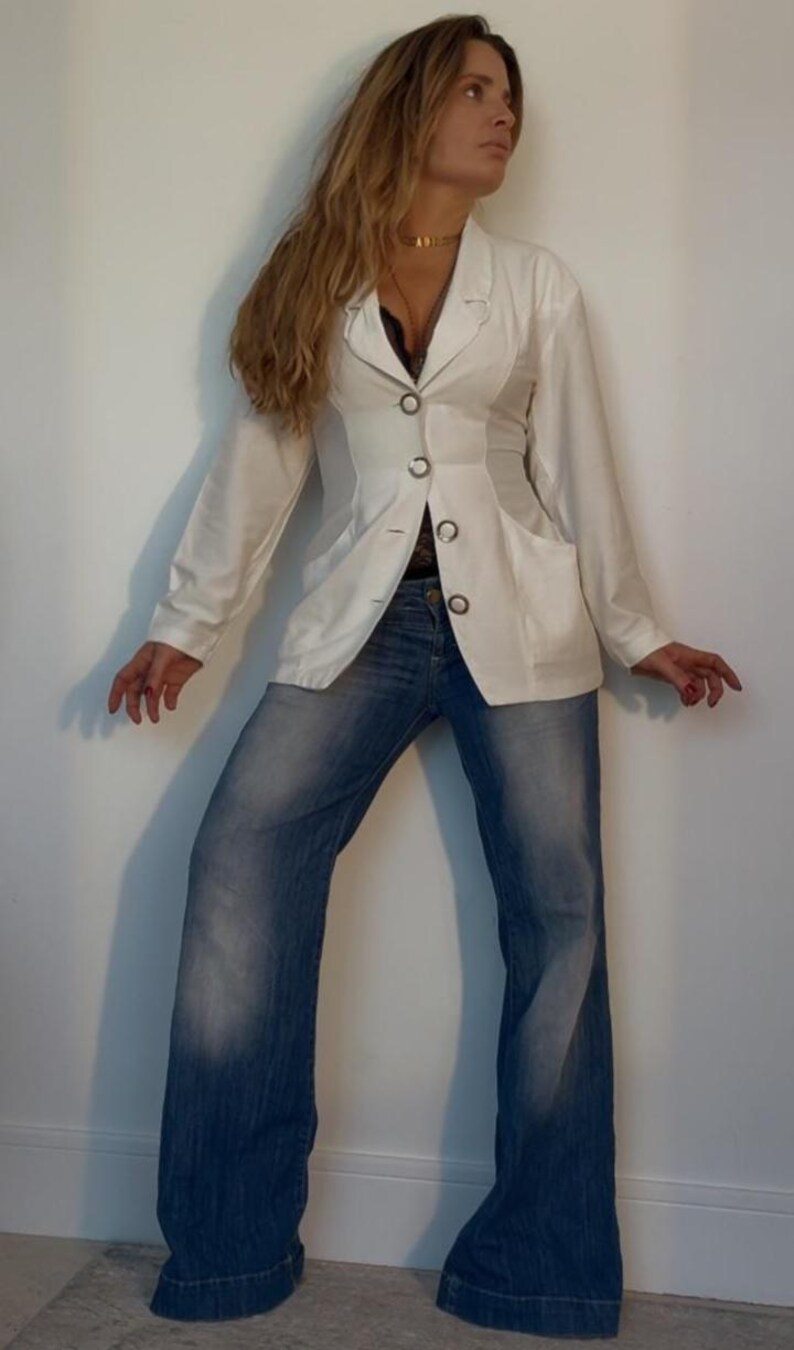 Puede incluir: Una blazer blanca con tres botones negros y dos bolsillos, combinada con vaqueros azules. La blazer tiene un corte entallado y los vaqueros tienen una pernera acampanada. El conjunto es sencillo y elegante, adecuado para diversas ocasiones.