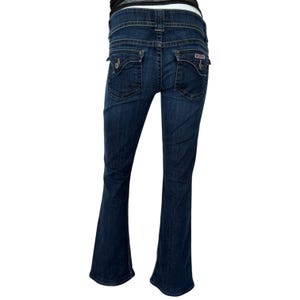 Może przedstawiać: Jeansy z ciemnego denimu z nogawką w kształcie dzwonu. Jeansy mają dwie tylne kieszenie z ozdobnymi przeszyciami i małą naszywką z logo. Klasyczny styl.