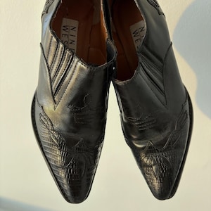 May include: A pair of black leather ankle boots with pointed toes. The boots feature decorative stitching and a textured pattern on the toe area. The brand name "NINE WEST" is visible on the inner lining.