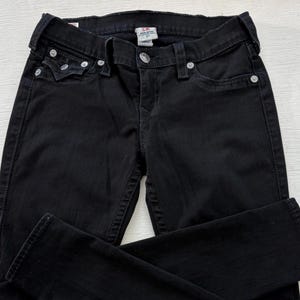 Puede incluir: Vaqueros de mezclilla negros con un dise&ntilde;o cl&aacute;sico de cinco bolsillos. Los vaqueros tienen remaches plateados y cierre de bot&oacute;n. La tela parece ser una mezclilla de peso medio, adecuada para el uso diario. Los vaqueros est&aacute;n doblados, mostrando el frente y parte de la espalda.