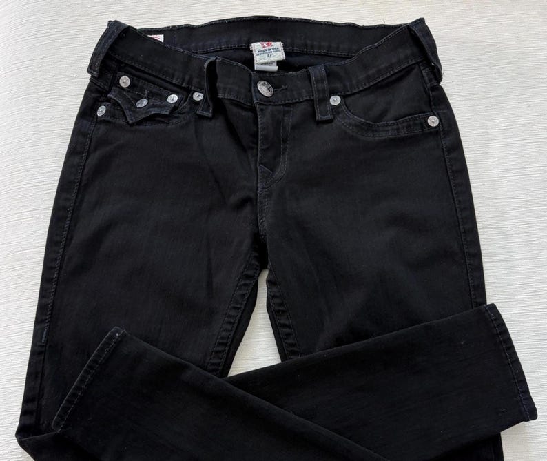 Puede incluir: Vaqueros de mezclilla negros con un dise&ntilde;o cl&aacute;sico de cinco bolsillos. Los vaqueros tienen remaches plateados y cierre de bot&oacute;n. La tela parece ser una mezclilla de peso medio, adecuada para el uso diario. Los vaqueros est&aacute;n doblados, mostrando el frente y parte de la espalda.