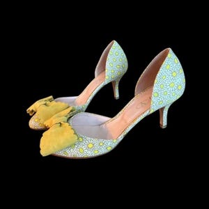 Op de afbeelding: Een paar lichtgroene en witte pumps met madeliefjespatroon en gele strik. De schoenen hebben een spitse neus en een kleine hak. De merknaam is zichtbaar op de binnenzool.