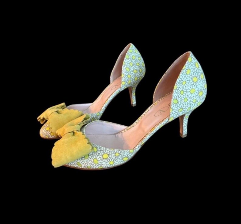 Op de afbeelding: Een paar lichtgroene en witte pumps met madeliefjespatroon en gele strik. De schoenen hebben een spitse neus en een kleine hak. De merknaam is zichtbaar op de binnenzool.