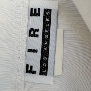 Puede incluir: Primer plano de una etiqueta de ropa blanca con la palabra "FIRE" en letras negras en negrita apiladas verticalmente. Debajo, las palabras "LOS ANGELES" est&aacute;n impresas verticalmente en una columna negra. La etiqueta est&aacute; adherida a tela blanca.