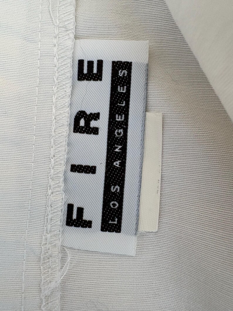 Puede incluir: Primer plano de una etiqueta de ropa blanca con la palabra "FIRE" en letras negras en negrita apiladas verticalmente. Debajo, las palabras "LOS ANGELES" est&aacute;n impresas verticalmente en una columna negra. La etiqueta est&aacute; adherida a tela blanca.