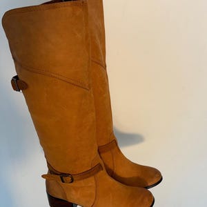 May include: A pair of knee-high, tan leather boots. The boots have a rounded toe, a small heel, and a strap with a buckle around the ankle. The leather has a slightly distressed appearance.