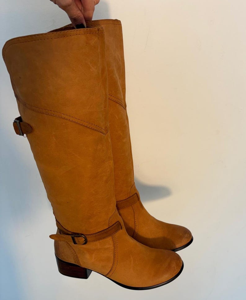 May include: A pair of knee-high, tan leather boots. The boots have a rounded toe, a small heel, and a strap with a buckle around the ankle. The leather has a slightly distressed appearance.