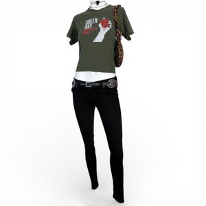 Puede incluir: Una camiseta verde oliva con el texto "Green Day American Idiot" y un gr&aacute;fico de una mano sosteniendo un coraz&oacute;n. El atuendo incluye unos vaqueros pitillo negros, un cintur&oacute;n negro con hebillas plateadas y un bolso de hombro con estampado de cebra.