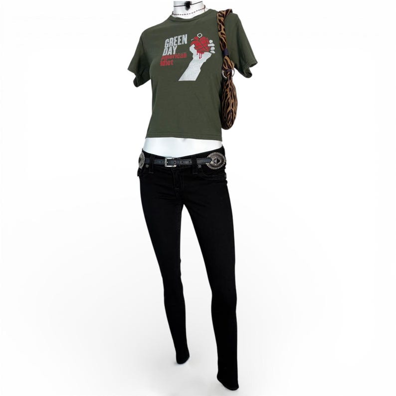 Puede incluir: Una camiseta verde oliva con el texto "Green Day American Idiot" y un gr&aacute;fico de una mano sosteniendo un coraz&oacute;n. El atuendo incluye unos vaqueros pitillo negros, un cintur&oacute;n negro con hebillas plateadas y un bolso de hombro con estampado de cebra.