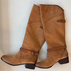 May include: A pair of tan leather knee-high boots. The boots have a rounded toe, a small heel, and adjustable straps with buckles. The leather has a slightly distressed appearance, and the boots are set against a neutral background.