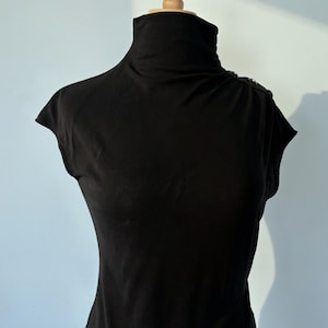 Puede incluir: Una blusa negra sin mangas con cuello alto y una hilera de pequeños botones redondos de tono metálico a lo largo de la costura del hombro izquierdo. La prenda se exhibe en un maniquí, mostrando una silueta ajustada y tela lisa.