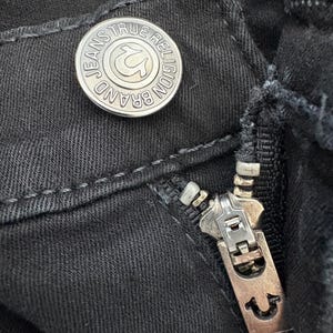 Puede incluir: Primer plano de unos vaqueros negros con un bot&oacute;n y una cremallera plateados. El bot&oacute;n lleva grabadas las palabras "JEANS TRUE RELIGION BRAND" alrededor de un logotipo de herradura. La cremallera tambi&eacute;n es plateada.