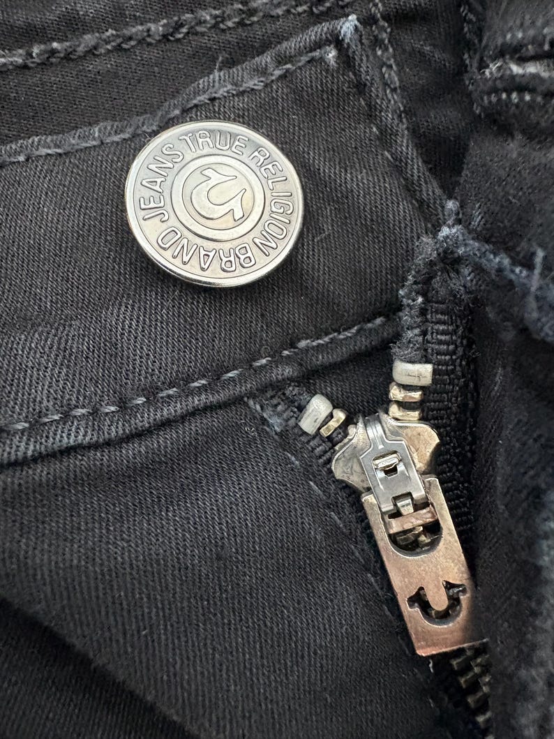 Puede incluir: Primer plano de unos vaqueros negros con un bot&oacute;n y una cremallera plateados. El bot&oacute;n lleva grabadas las palabras "JEANS TRUE RELIGION BRAND" alrededor de un logotipo de herradura. La cremallera tambi&eacute;n es plateada.