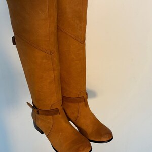 May include: A pair of tall, tan leather boots. The boots have a knee-high design with a rounded toe and a small heel. They feature a strap and buckle detail around the ankle and a decorative stitch pattern. The leather has a slightly distressed appearance.