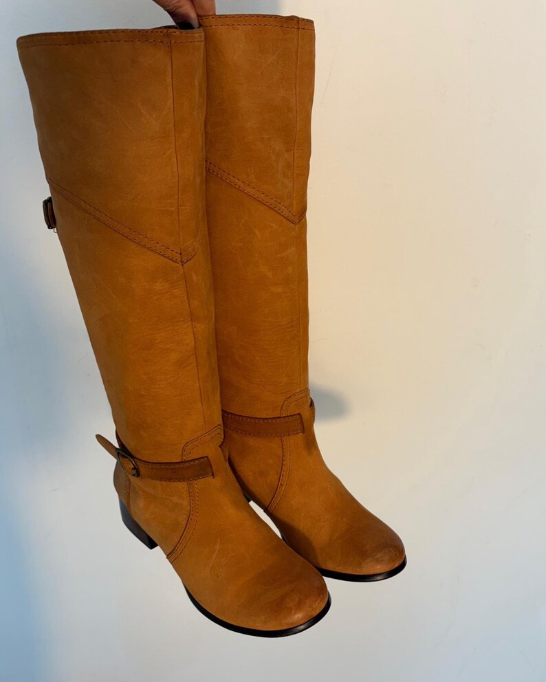 May include: A pair of tall, tan leather boots. The boots have a knee-high design with a rounded toe and a small heel. They feature a strap and buckle detail around the ankle and a decorative stitch pattern. The leather has a slightly distressed appearance.