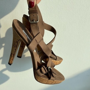 Puede incluir: Sandalias marrones de tacón alto con plataformas y tacones de corcho. Las sandalias tienen múltiples correas y hebillas, con una altura de tacón de aproximadamente 10 cm.