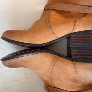 May include: Tan leather boots with a stacked heel and a strap detail. The boots have a rounded toe and a dark sole. The leather has a distressed look, and the boots appear to be well-worn.