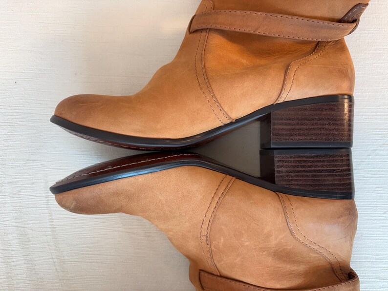 May include: Tan leather boots with a stacked heel and a strap detail. The boots have a rounded toe and a dark sole. The leather has a distressed look, and the boots appear to be well-worn.