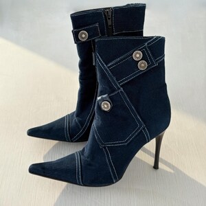 May include: A pair of dark navy blue corduroy ankle boots with a pointed toe and a high stiletto heel. The boots feature white contrast stitching, decorative silver-toned metal buttons, and a side zipper closure on the inner shaft of each boot.