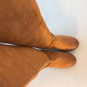 May include: A pair of knee-high, tan leather boots. The boots have a rounded toe and a smooth, matte finish. The leather appears to be slightly creased, adding texture to the design. The boots are set against a white background.