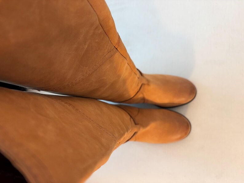 May include: A pair of knee-high, tan leather boots. The boots have a rounded toe and a smooth, matte finish. The leather appears to be slightly creased, adding texture to the design. The boots are set against a white background.