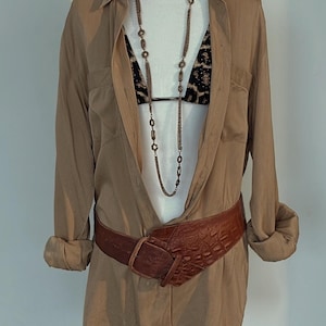 Puede incluir: Una camisa de manga larga color canela con botones y una sutil textura acanalada vertical, sobre un top de bikini con estampado de leopardo negro y canela. Un collar largo de cadena color bronce con eslabones circulares y un cinturón ancho marrón.