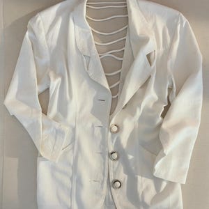 Puede incluir: Un blazer blanco con un dise&ntilde;o &uacute;nico de espalda abierta. El blazer presenta una solapa con muescas cl&aacute;sica, cierre de tres botones y dos bolsillos delanteros. La espalda tiene una serie de correas blancas. La prenda est&aacute; hecha de tela ligera.