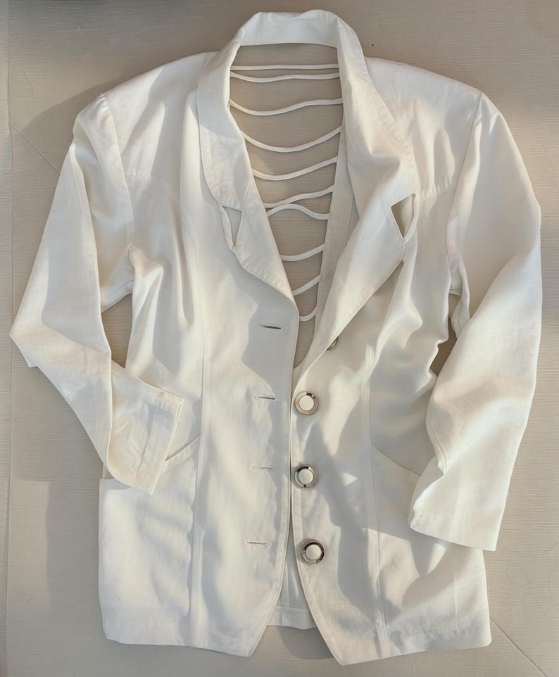 Puede incluir: Un blazer blanco con un dise&ntilde;o &uacute;nico de espalda abierta. El blazer presenta una solapa con muescas cl&aacute;sica, cierre de tres botones y dos bolsillos delanteros. La espalda tiene una serie de correas blancas. La prenda est&aacute; hecha de tela ligera.