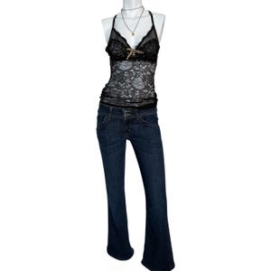 Może przedstawiać: Czarny top z koronki z złotym akcentem i ciemne dżinsy. Jeansy mają niski stan i rozszerzane nogawki. Widoczny jest r&oacute;wnież naszyjnik z małą zawieszką.