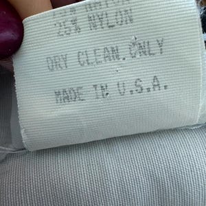 Puede incluir: Primer plano de una etiqueta de tela blanca con el texto "25% NYLON, DRY CLEAN ONLY, MADE IN U.S.A.". La etiqueta est&aacute; sobre una tela azul claro con textura acanalada.