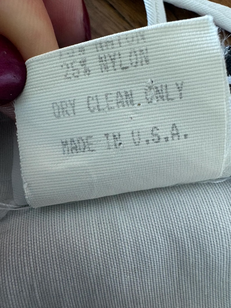 Puede incluir: Primer plano de una etiqueta de tela blanca con el texto "25% NYLON, DRY CLEAN ONLY, MADE IN U.S.A.". La etiqueta est&aacute; sobre una tela azul claro con textura acanalada.