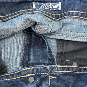 Może przedstawiać: Zbliżenie na parę niebieskich dżins&oacute;w. Obraz przedstawia pas, szwy i metalowy guzik. Nazwa marki "HUDSON" jest widoczna na etykiecie.