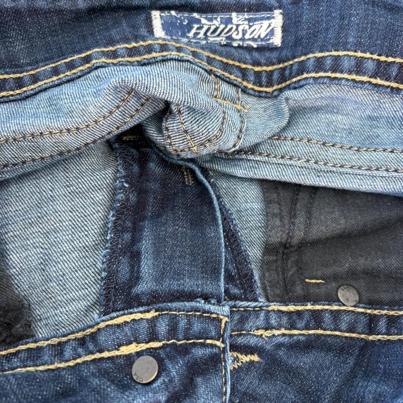 Może przedstawiać: Zbliżenie na parę niebieskich dżins&oacute;w. Obraz przedstawia pas, szwy i metalowy guzik. Nazwa marki "HUDSON" jest widoczna na etykiecie.