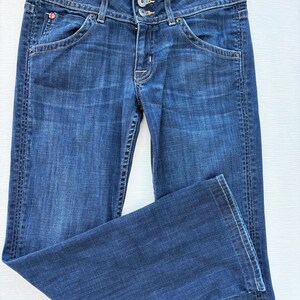 Może przedstawiać: Ciemnoniebieskie dżinsy z klasycznym pięciokieszeniowym designem. Dżinsy mają zapięcie na guzik i zamek błyskawiczny, szlufki na pasek i markę "Hudson" na pasku. Dżinsy są złożone, prezentując fakturę i pranie denimu.